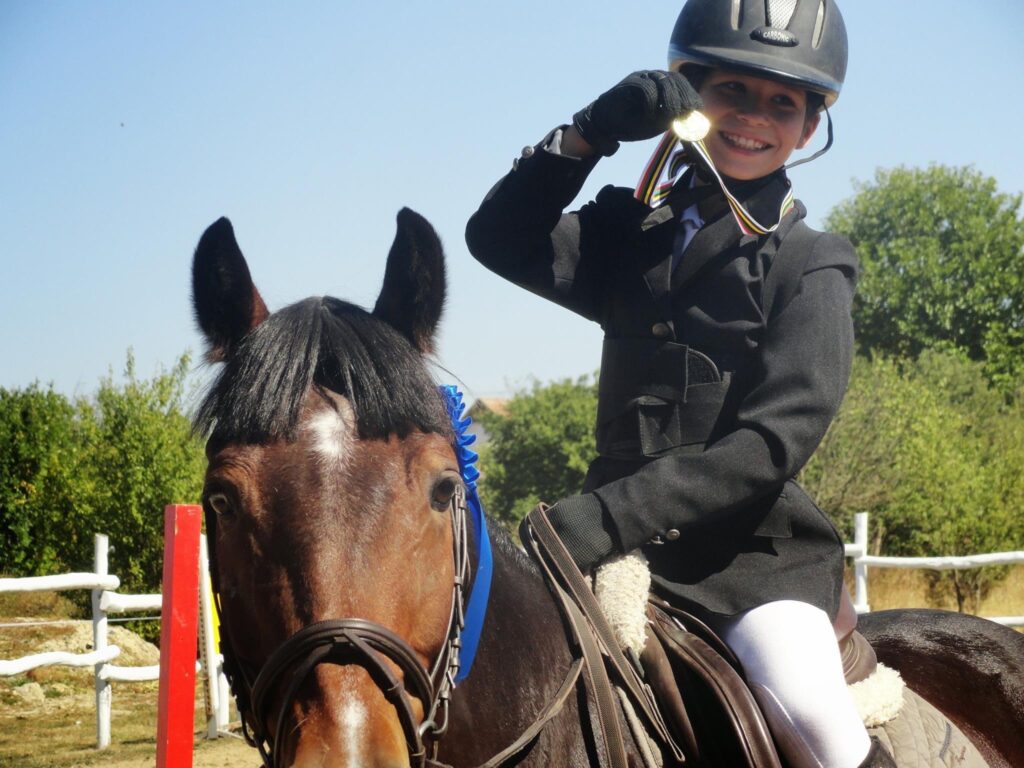 Egy fiatal lovas aranyérmet mutogat mosolyogva a lova hátán ülve egy versenyen